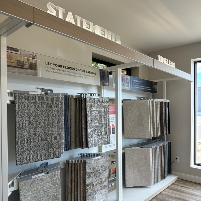 Carpet showroom display with 'STATEMENTS' and 'TIMELESS' sections, featuring hanging carpet samples in various patterns and colors, a 'RUGMASTER' protector sign, and a wooden floor.